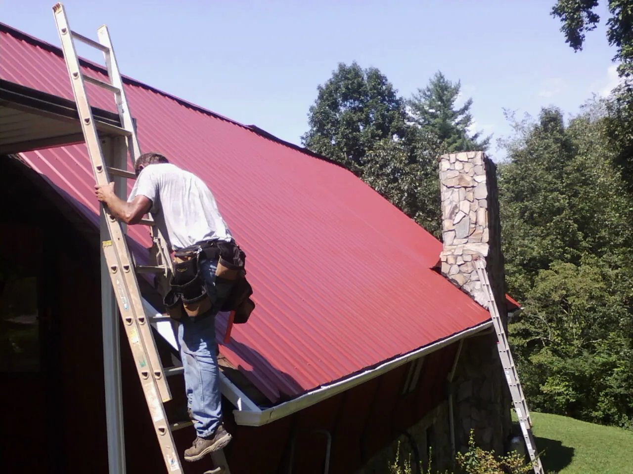 Roofer on a ladder beside a red metal roof and stone chimney — Soffit & Fascia Repair by Roofing Brothers in New Deal